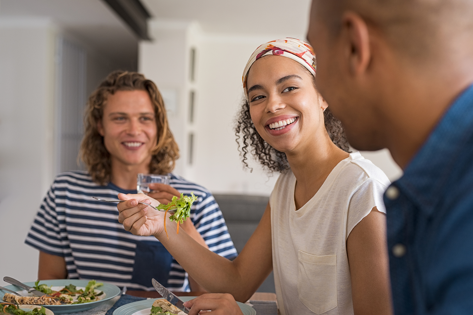 Beautiful african woman eating fresh salad with friends for lunch. Happy cheerful friends in a conversation during lunch at home. Young friends meeting each other for healthy meal.